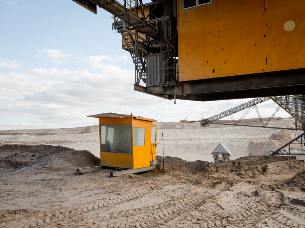 Tagebau Jänschwalde
Residuum 
Ende 2023 wurde im Tagebau Jänschwalde planmäßig die Kohleförderung eingestellt. Nach der Sicherung der Flöz- und Kippenflächen begann unmittelbar die Rekultivierung des ehemaligen Bergbaugebiets. In der riesigen Grube werden Sand- und Erdmassen stabilisiert, damit künftig Wälder, landwirtschaftliche Flächen und eine neue Seenlandschaft entstehen können. Rund 130 Bergleute sind noch mit diesen Arbeiten beschäftigt. Ina Schoenenburg fotografierte sie in dieser kargen Landschaft, an ihrem (letzten) Arbeitsplatz mit seinen Maschinen von beeindruckender Größe.